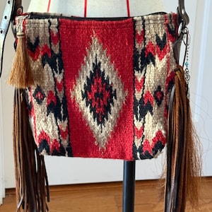 May include: A crossbody bag with a woven red, black, and beige geometric pattern. The bag features brown leather fringe and a matching leather strap. A small tassel hangs from the zipper.