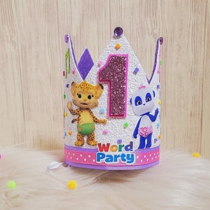 Puede incluir: Una corona blanca con un n&uacute;mero uno rosa, ribete morado y confeti de colores. La corona presenta im&aacute;genes de un guepardo y un panda. La corona tambi&eacute;n tiene las palabras "Word Party" en la parte inferior.