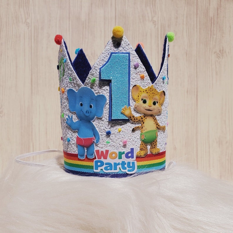 Puede incluir: Una corona de cumplea&ntilde;os azul y blanca con un n&uacute;mero uno, un elefante azul y un guepardo amarillo. La corona tiene una franja de arco&iacute;ris y las palabras "Word Party" en ella.