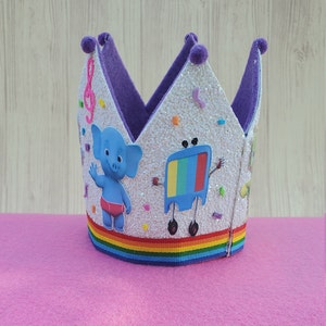 Puede incluir: Una corona blanca de purpurina con forro de fieltro morado y cinta de arco&iacute;ris. La corona presenta un elefante de dibujos animados y una televisi&oacute;n con cara.
