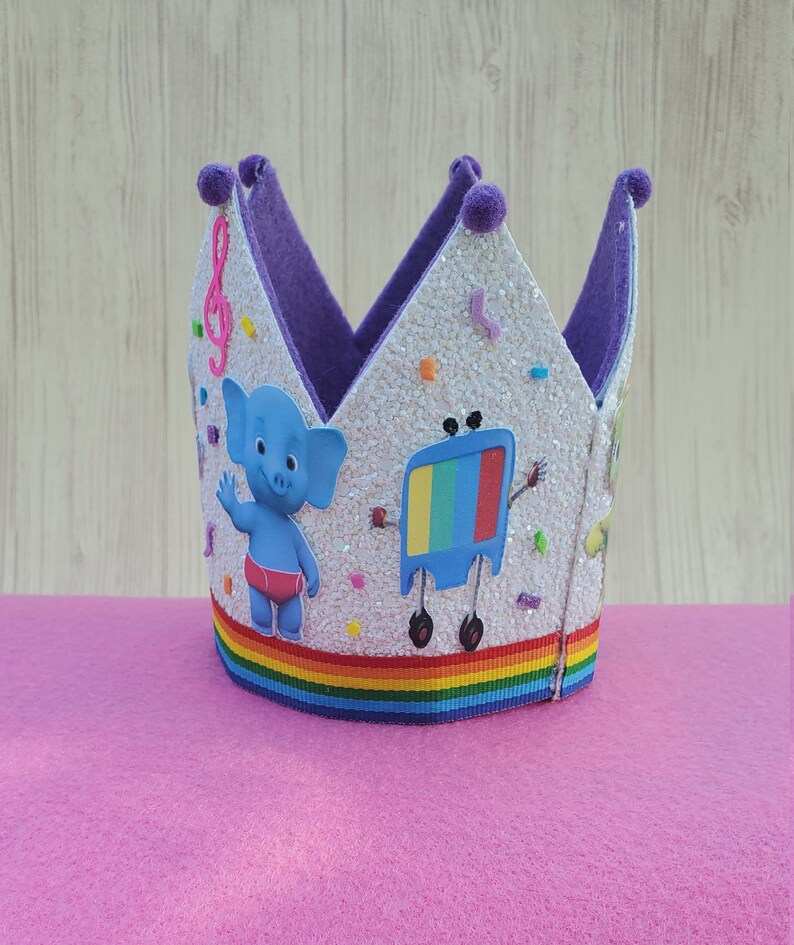 Puede incluir: Una corona blanca de purpurina con forro de fieltro morado y cinta de arco&iacute;ris. La corona presenta un elefante de dibujos animados y una televisi&oacute;n con cara.