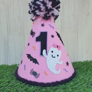 K&ouml;nnte beinhalten: Rosa Filzhut mit einem schwarzen und rosa Bommel oben. Der Hut ist mit einer schwarzen Filznummer eins, einem wei&szlig;en Filzgespenst mit Glitzer, schwarzen Filzflederm&auml;usen, schwarzen Filzspinnen und orangefarbenen und violetten Filzkonfetti verziert.