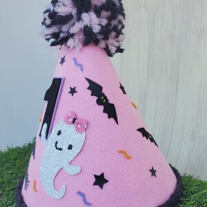 K&ouml;nnte beinhalten: Rosa Filzhut mit einem schwarzen Bommel oben. Der Hut ist mit einem Geist, Flederm&auml;usen, Sternen und Zickzacklinien verziert. Die Zahl "1" befindet sich an der Seite des Hutes.