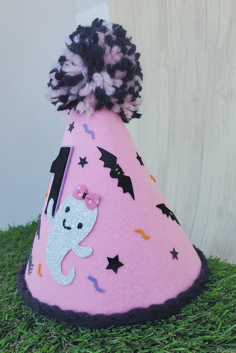 K&ouml;nnte beinhalten: Rosa Filzhut mit einem schwarzen Bommel oben. Der Hut ist mit einem Geist, Flederm&auml;usen, Sternen und Zickzacklinien verziert. Die Zahl "1" befindet sich an der Seite des Hutes.