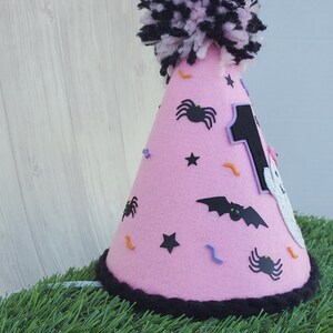 K&ouml;nnte beinhalten: Rosa Filzhut mit einem schwarzen Bommel oben. Der Hut ist mit schwarzen Spinnen, Flederm&auml;usen, Sternen und orangefarbenen und violetten Konfetti verziert. Die Zahl "1" befindet sich auf der Vorderseite des Hutes.
