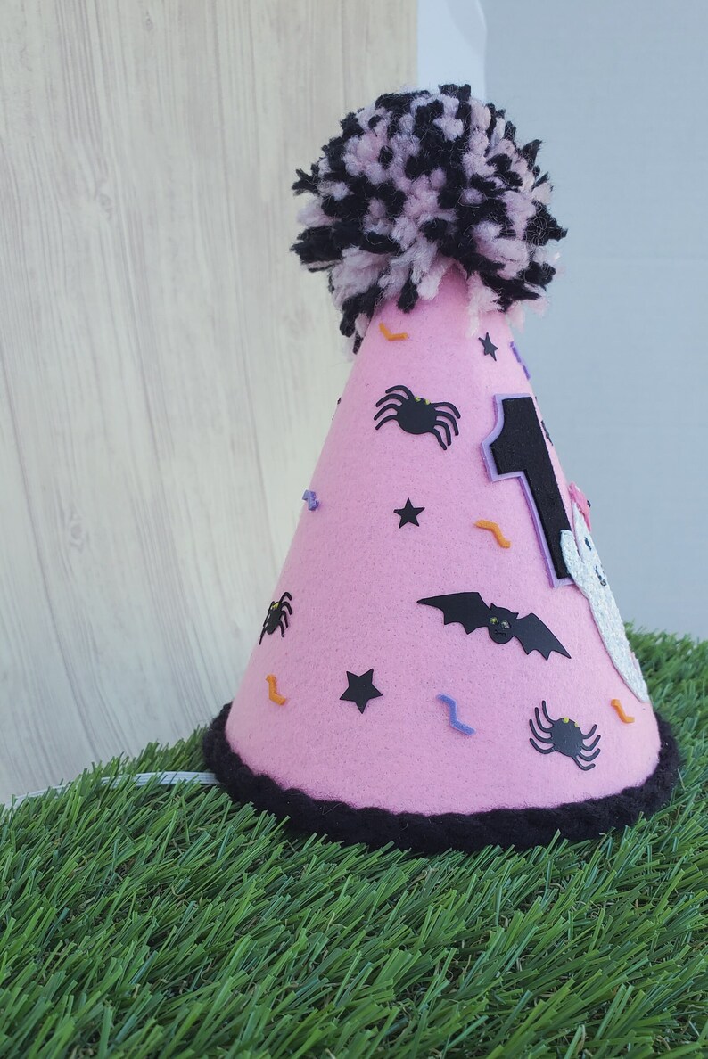K&ouml;nnte beinhalten: Rosa Filzhut mit einem schwarzen Bommel oben. Der Hut ist mit schwarzen Spinnen, Flederm&auml;usen, Sternen und orangefarbenen und violetten Konfetti verziert. Die Zahl "1" befindet sich auf der Vorderseite des Hutes.
