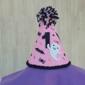 K&ouml;nnte beinhalten: Rosa Filzhut mit einem schwarz-wei&szlig;en Bommel oben. Der Hut hat eine schwarze Filznummer eins, einen wei&szlig;en Geist mit einer rosa Schleife, schwarze Flederm&auml;use, Spinnen und Konfetti.