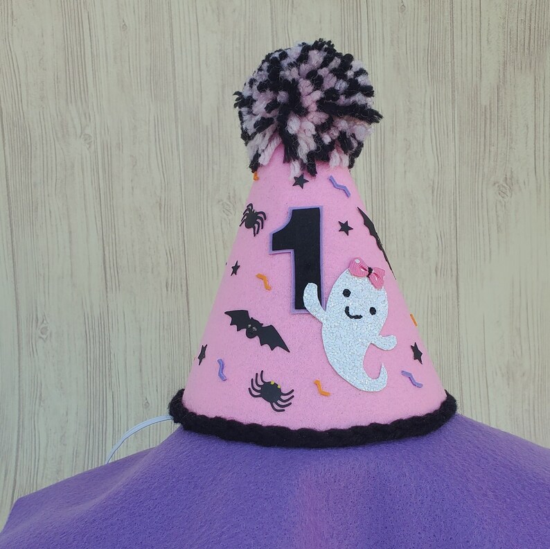 K&ouml;nnte beinhalten: Rosa Filzhut mit einem schwarz-wei&szlig;en Bommel oben. Der Hut hat eine schwarze Filznummer eins, einen wei&szlig;en Geist mit einer rosa Schleife, schwarze Flederm&auml;use, Spinnen und Konfetti.