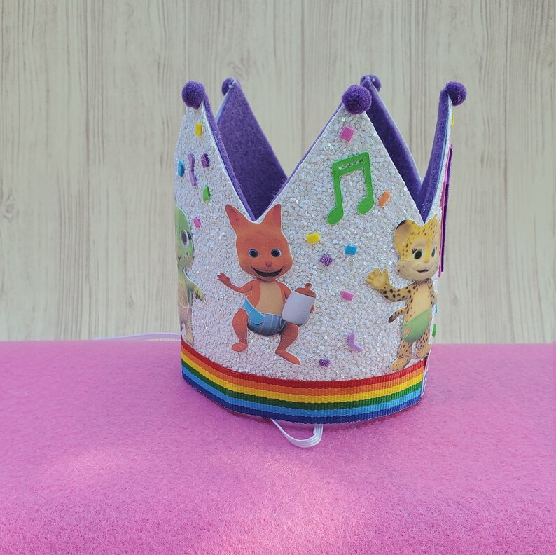 Puede incluir: Corona de cumplea&ntilde;os morada y blanca con ribete de arco&iacute;ris. La corona presenta una imagen de dibujos animados de tres personajes del programa infantil "Yo Gabba Gabba!" Los personajes son un monstruo verde, un canguro rojo y un guepardo amarillo.