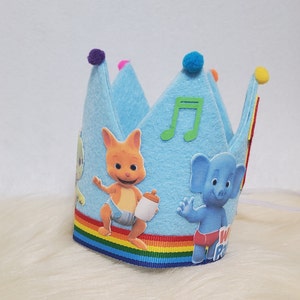 May include: Blue felt crown with a rainbow trim. The crown features a musical note, a kangaroo character wearing a nappy and holding a bottle, and a blue elephant character wearing a red nappy. The text "We" is visible on the elephant's nappy.