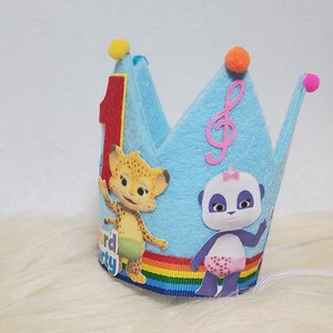 May include: Blue felt crown with a rainbow ribbon, featuring a red number one, a musical note, and two cartoon characters: a cheetah and a panda. The crown says "Word Party" on the bottom.