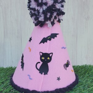 K&ouml;nnte beinhalten: Rosa Filzhut mit einem schwarzen Bommel oben. Der Hut ist mit schwarzen Flederm&auml;usen, einer schwarzen Katze, einer Spinne und Sternen verziert.