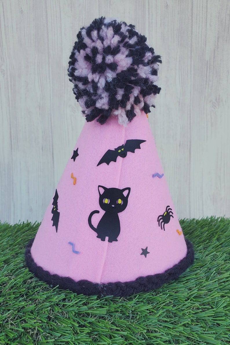 K&ouml;nnte beinhalten: Rosa Filzhut mit einem schwarzen Bommel oben. Der Hut ist mit schwarzen Flederm&auml;usen, einer schwarzen Katze, einer Spinne und Sternen verziert.