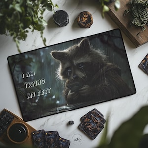 May include: A rectangular mousepad features a raccoon holding cards in the rain with the text "I AM TRYING MY BEST". The mousepad is surrounded by playing cards, a wooden coaster, and decorative stones. The color palette includes shades of brown, gray, and white.
