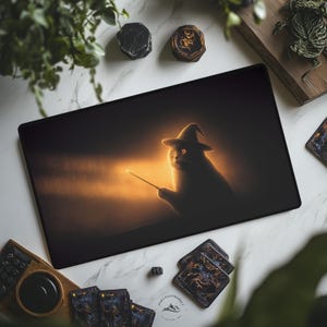 May include: A black rectangular desk mat featuring a cat wearing a wizard hat and holding a wand. The cat is silhouetted against a warm, glowing background. The mat is surrounded by other items, including playing cards and decorative stones.