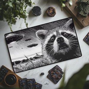 May include: Black and white mousepad featuring a detailed illustration of a raccoon looking up at UFOs in the sky. The design includes a field of wheat and the Paw Coast Market logo.