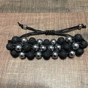 May include: A bracelet featuring black lava stone and silver beads. The design includes two rows of black, textured lava stone beads and a central row of shiny silver beads. The adjustable black cord allows for a comfortable fit.