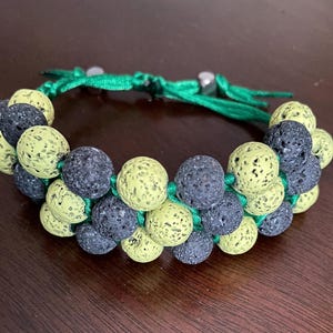 Könnte beinhalten: Ein handgefertigtes Armband mit abwechselnden Reihen aus schwarzen und hellgrünen Lavasteinperlen. Die Perlen sind auf einer grünen Kordel aufgefädelt, und das Armband hat einen Schiebeknotenverschluss. Das Armband ist verstellbar.