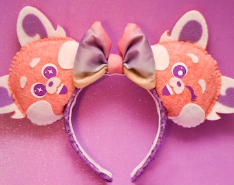 May include: A pink headband with a rainbow-colored bow and two plush ears featuring a cartoon character with a pink face, white spots, and purple eyes.