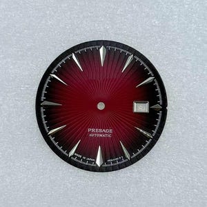 Könnte beinhalten: Ein Zifferblatt mit einem tiefroten Zentrum und einem schwarzen Außenring. Das Zifferblatt hat silberfarbene Markierungen und Zeiger mit dem Text "PRESAGE AUTOMATIC" und "MADE IN JAPAN". Das Zifferblatt hat ein Datumsfenster bei 3 Uhr.