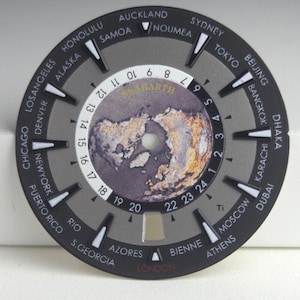 Puede incluir: Una esfera de reloj circular con un gráfico de mapa mundial en el centro y el texto SEABARTH. El anillo exterior muestra nombres de ciudades y una escala de 24 horas con marcadores blancos. La esfera tiene un fondo gris y ventana de fecha.