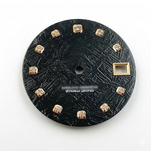 May include: A black watch face with a textured surface and gold-colored square markers. The face has a small hole in the center and the text "OFFICIALLY CERTIFIED" at the bottom. The markers are evenly spaced around the perimeter.