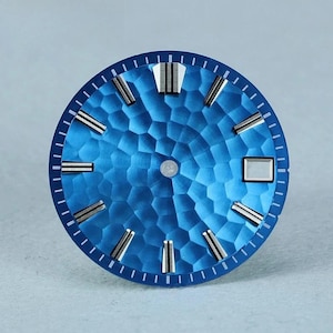 Pode incluir: Um mostrador de relógio circular com um acabamento azul vibrante e um padrão de favo de mel martelado. O mostrador inclui marcadores de hora aplicados em tom prateado com linhas centrais pretas e uma janela de data retangular às três.
