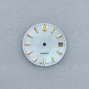 May include: A watch face with a pearlescent background and gold-colored hour markers. The face has the word "AUTOMATIC" printed at the bottom, with "JAPAN MOVT" below it. The face has a small square window for the date.