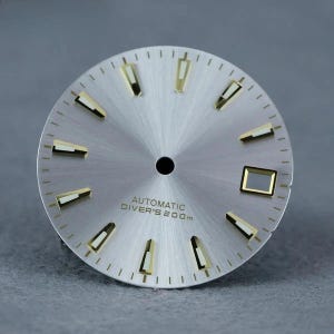 Puede incluir: Una esfera de reloj circular plateada con acabado rayos de sol, con marcadores de hora aplicados en tono dorado con centros luminosos blancos. El texto AUTOMATIC DIVER'S 200m está impreso en negro bajo el orificio central, con ventana de fecha a las tres.