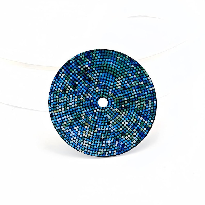 May include: Round decorative item with a mosaic design in blues, greens, and white. It has a central hole and a radial pattern of small, coloured tiles. The object is set against a white backdrop.