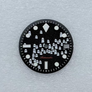 Puede incluir: Una esfera de reloj redonda y negra con una superficie texturizada y un patrón de pequeñas ilustraciones de fantasmas blancos. La esfera incluye marcadores de hora blancos circulares y rectangulares, un orificio central y la palabra Automatic en rojo.