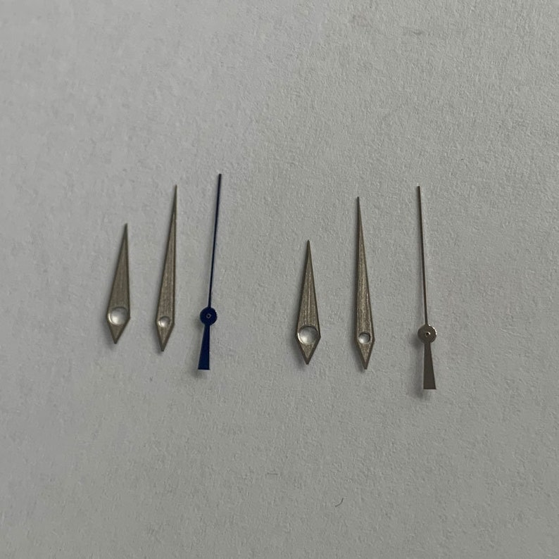 May include: A set of six watch hands, including a blue hour hand, a silver minute hand, a silver second hand, and three silver hands for a watch with a date complication.