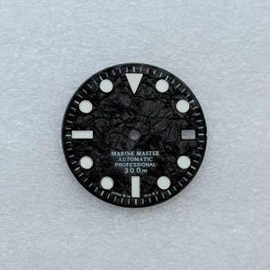 Könnte beinhalten: Schwarzes Zifferblatt mit weißen Stundenmarkierungen und Zeigern. Auf dem Zifferblatt steht "MARINE MASTER AUTOMATIC PROFESSIONAL 300m". Das Zifferblatt hat eine strukturierte Oberfläche und ein weißes Dreieck bei 12 Uhr.