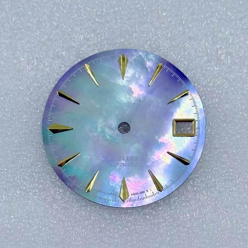 Puede incluir: Una esfera de reloj redonda con un acabado nacarado e iridiscente. La esfera tiene marcadores de hora triangulares de color dorado y una peque&ntilde;a ventana rectangular. El texto "PRESAGE AUTOMATIC" est&aacute; impreso en la esfera.