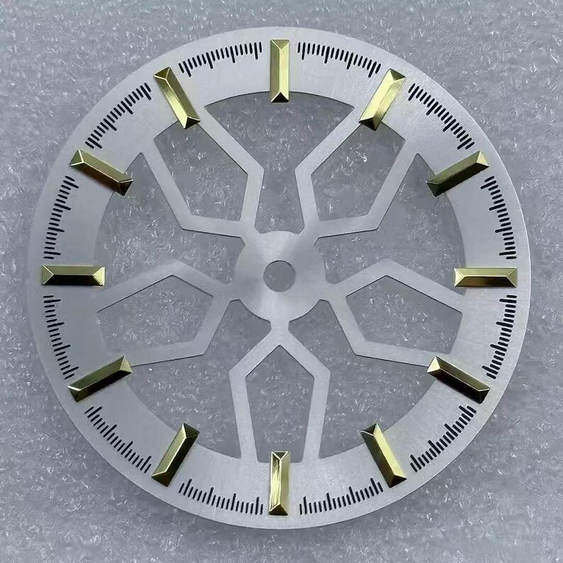 Pode incluir: Um mostrador de rel&oacute;gio prateado com marcadores de hora retangulares dourados. O mostrador tem um design geom&eacute;trico no centro e uma escala de minutos ao redor do per&iacute;metro. Este mostrador &eacute; um componente para a relojoaria, com cerca de 3,5 cm de di&acirc;metro.