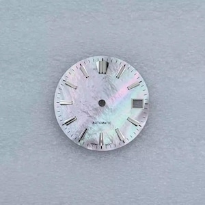 May include: A watch face with a pearlescent, iridescent finish. The face has silver-toned hour markers and hands. The word "AUTOMATIC" is printed at the bottom. The watch face has a date window and is marked "JAPAN MOVT".