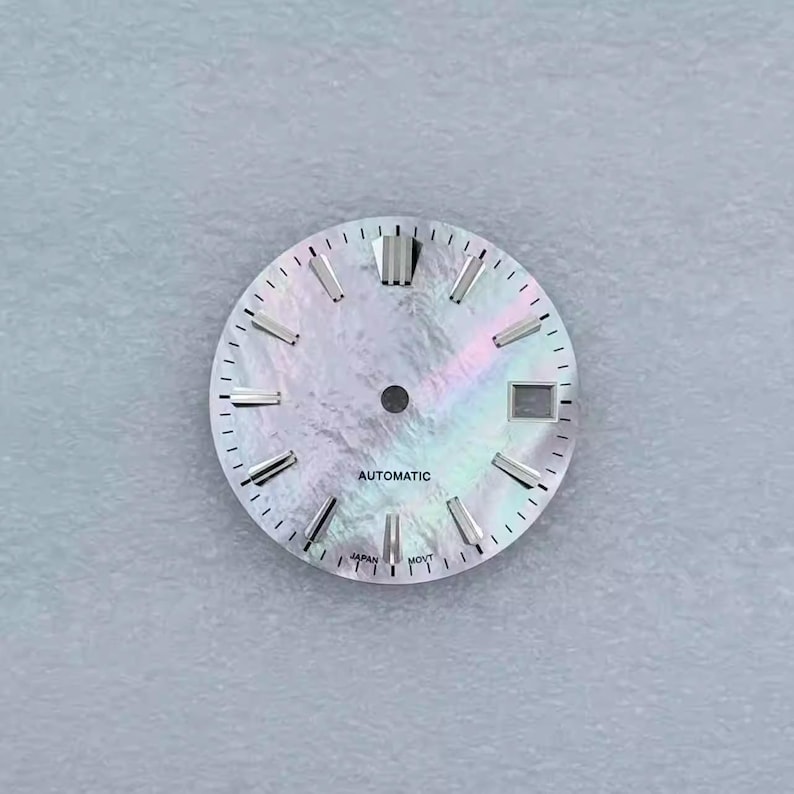 May include: A watch face with a pearlescent, iridescent finish. The face has silver-toned hour markers and hands. The word "AUTOMATIC" is printed at the bottom. The watch face has a date window and is marked "JAPAN MOVT".