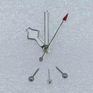 May include: A collection of watch hands and components. The hands are silver-toned with a red-tipped second hand. The hour and minute hands have white accents. Several small, silver-toned watch hands are also present, likely for other functions.
