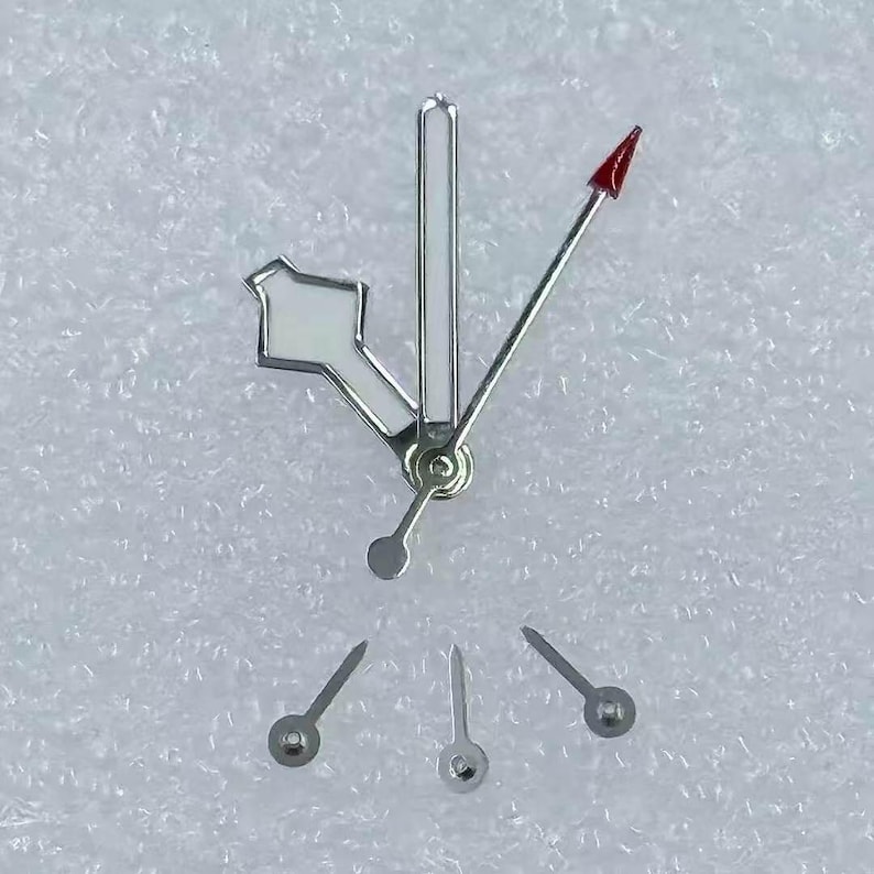 May include: A collection of watch hands and components. The hands are silver-toned with a red-tipped second hand. The hour and minute hands have white accents. Several small, silver-toned watch hands are also present, likely for other functions.