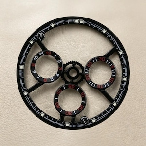 Puede incluir: Un componente de esfera de reloj circular negro con un diseño esqueletizado y tres subesferas. El anillo exterior muestra marcadores de minutos blancos e índices cuadrados. Las subesferas muestran números en rojo y blanco, con un engranaje central.