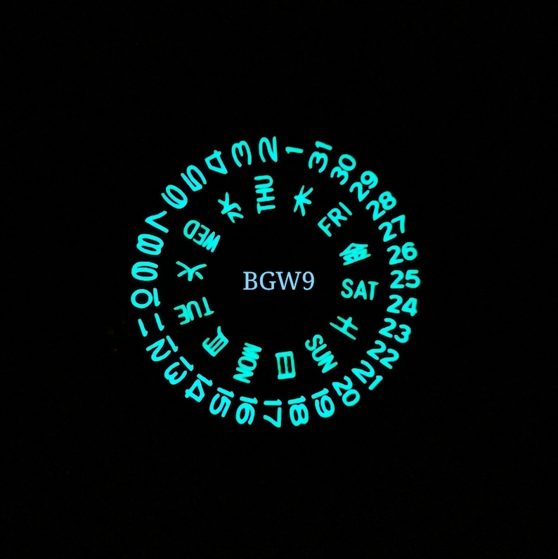 May include: A circular watch face with a black background and a glowing turquoise dial. The dial displays the days of the week, the numbers 1 through 31, and the text "BGW9".