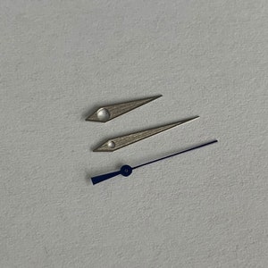 May include: Three watch hands, two silver and one blue. The silver hands are pointed and the blue hand is a thicker, flat shape.