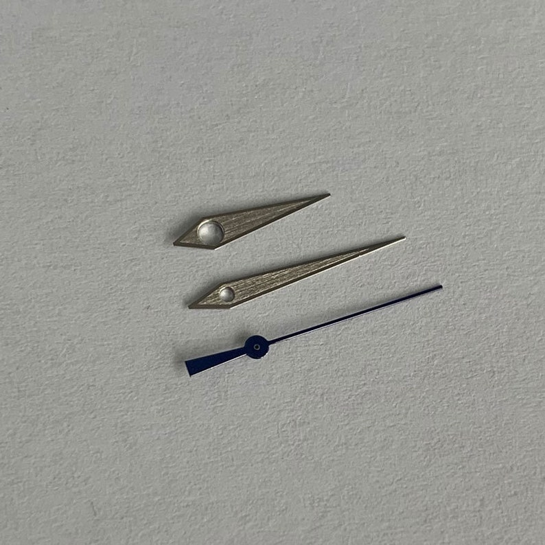 May include: Three watch hands, two silver and one blue. The silver hands are pointed and the blue hand is a thicker, flat shape.