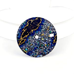 May include: A round watch face with a unique design. The face features a dark blue background with a pattern of small, colorful circles in shades of blue, green, yellow, and white. A gold-colored, abstract shape is in the center.