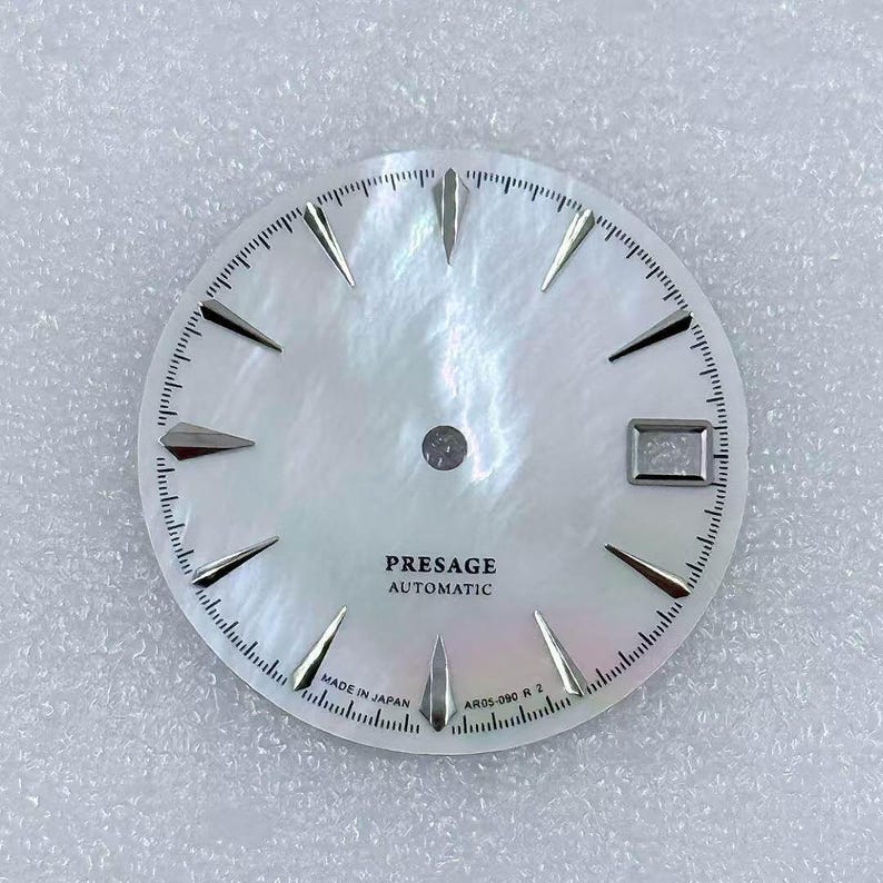 Puede incluir: Una esfera de reloj con una esfera blanca nacarada. La esfera presenta marcadores de hora plateados, una ventana de fecha y el texto "PRESAGE AUTOMATIC" y "MADE IN JAPAN". Tambi&eacute;n es visible el n&uacute;mero de modelo AR05-090 R 2.