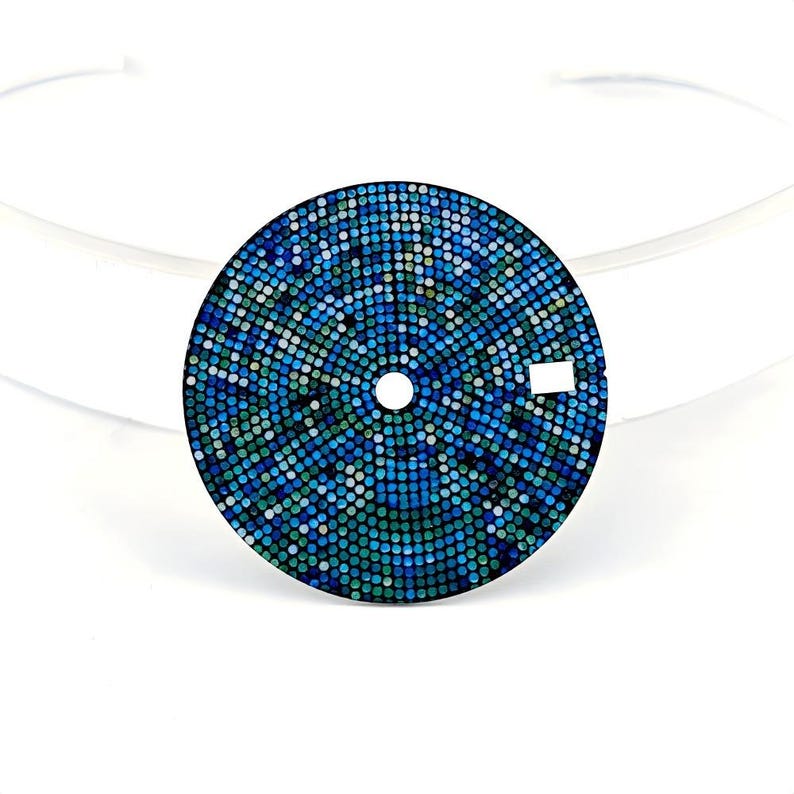 May include: A circular watch face featuring a mosaic design of blue and green squares. It has a central hole and a small rectangular marker. The background is white.