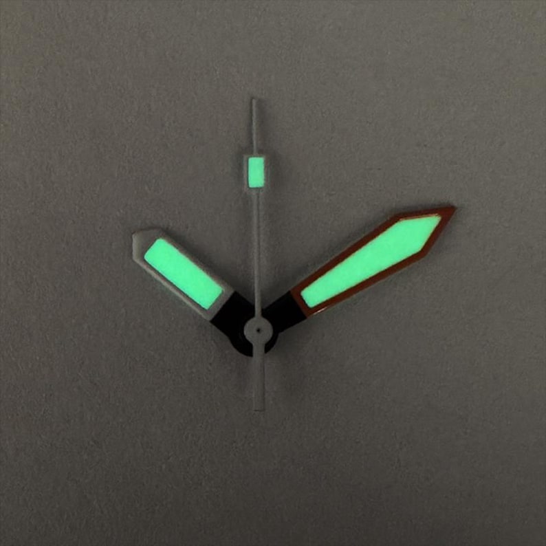 May include: Close-up of watch hands with a luminous green glow. The hour and minute hands are black with green rectangular and arrow-shaped tips. The second hand has a green rectangular tip. The hands are set against a gray background.