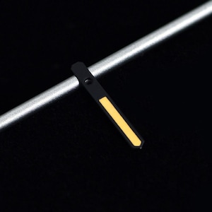 May include: Close-up of a black watch hand with a yellow stripe. The hand is positioned diagonally against a silver bar and a black background. The hand has a small hole near the top.