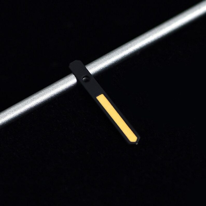 May include: Close-up of a black watch hand with a yellow stripe. The hand is positioned diagonally against a silver bar and a black background. The hand has a small hole near the top.