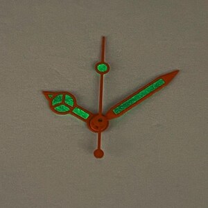 May include: Orange watch hands with glowing green accents. The hands are a vibrant orange color, with luminous green detailing. The design includes a second hand, minute hand, and hour hand, all with a unique, eye-catching style. The hands are designed for a watch.